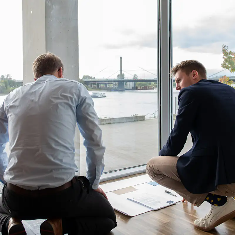 Zwei Seminarteilnehmer knien ihre Arbeit besprechend vor einer großen Festerfront mit dem Rhein in Leverkusen im Hintergrund